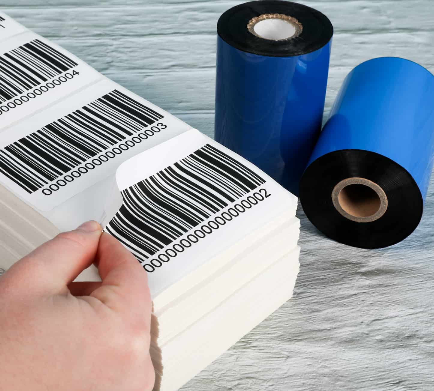 a hand peeling off a barcode label from a stack of fanfold labels with 2 thermal ribbons in the background
