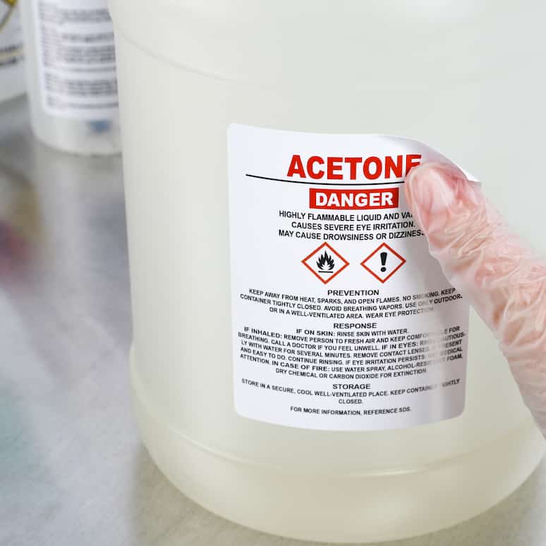 a gloved hand applying a chemical label to a jug of acetone on a metal table, in the background are similar jugs