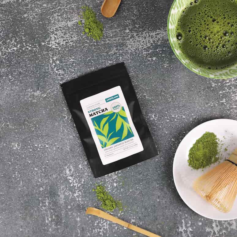 a packet of matcha sits surrounded by a small bamboo scoop, a bamboo whisk on a plate, and a cup of brewed matcha tea, all of the tools have a pilf of spilled matcha next to them