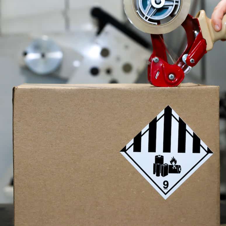 a hand uses a tape gun to seal a cardboard box with a battery danger label