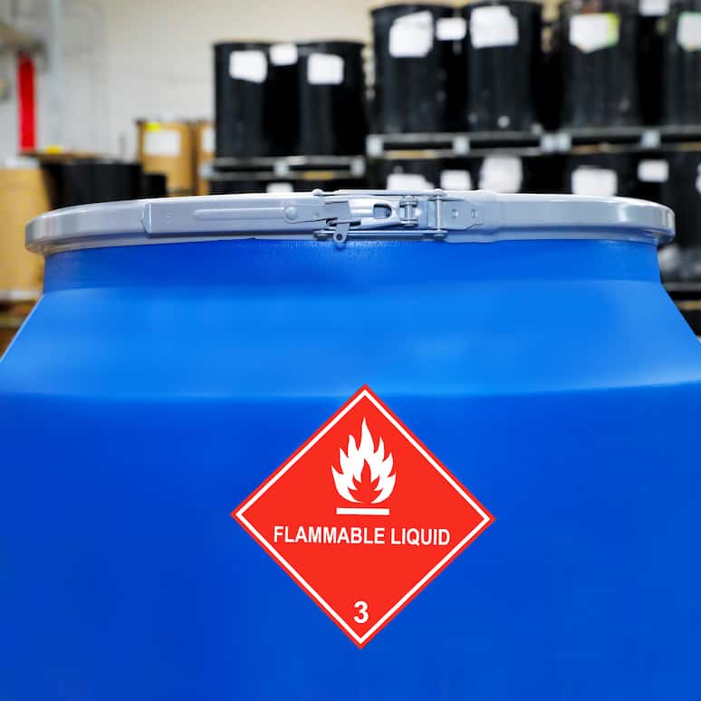 a large plastic barrel with a flammable gas label sits in a factory, in the backround are rows and rows of barrels stacked on pallettes