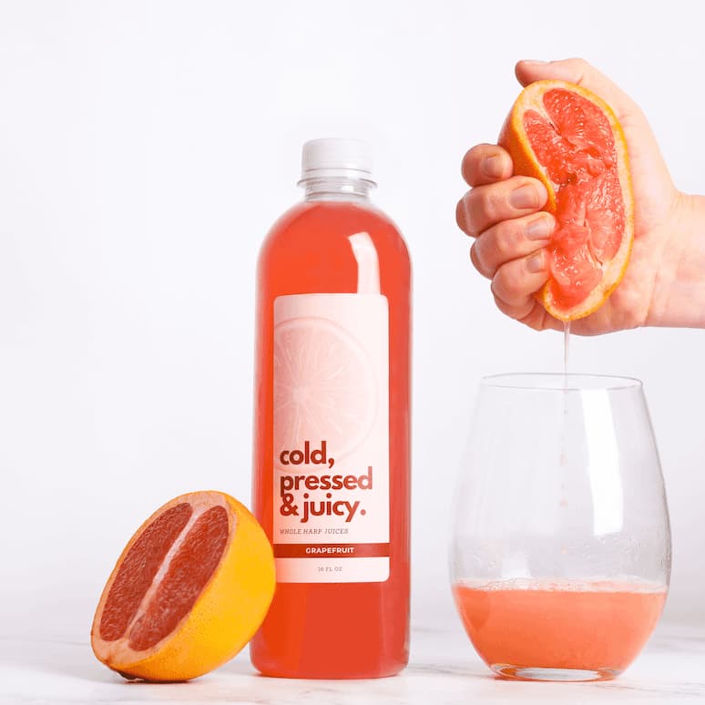 a bottle of grapefruit juice with a half grapefruit leaning against it, to the right a hand squeezes a grapefruit and the juice fills a glass sitting below