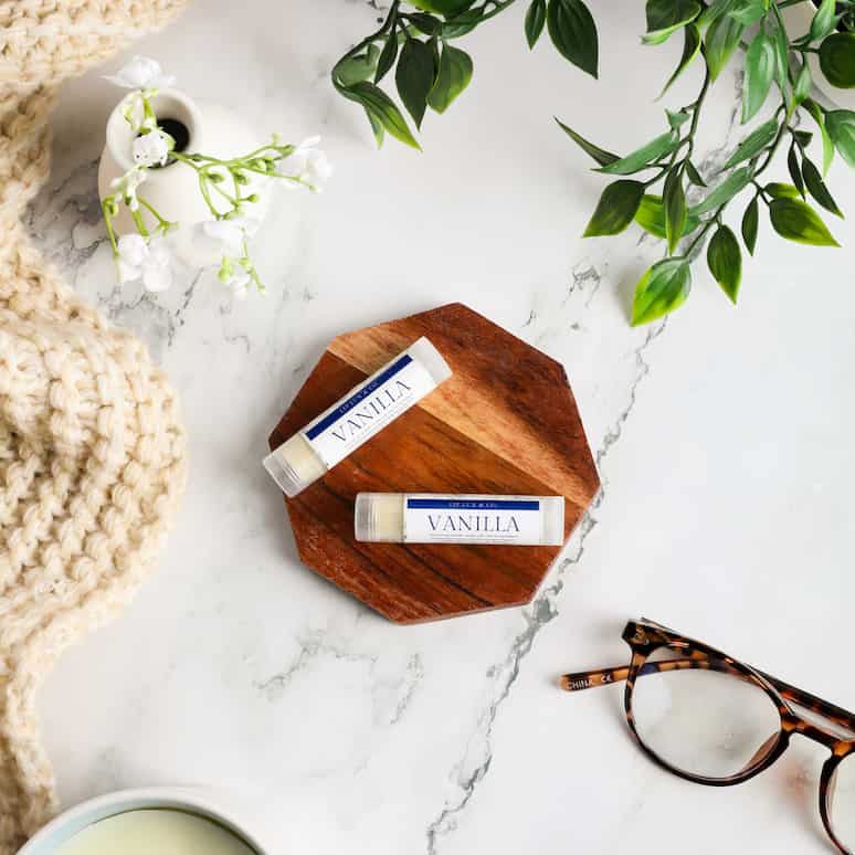 two vanilla lip balms sitting on a wooden coaster surrounded by a pair of glasses, a candle, a quilt, fake plants in a vase, and fake vines