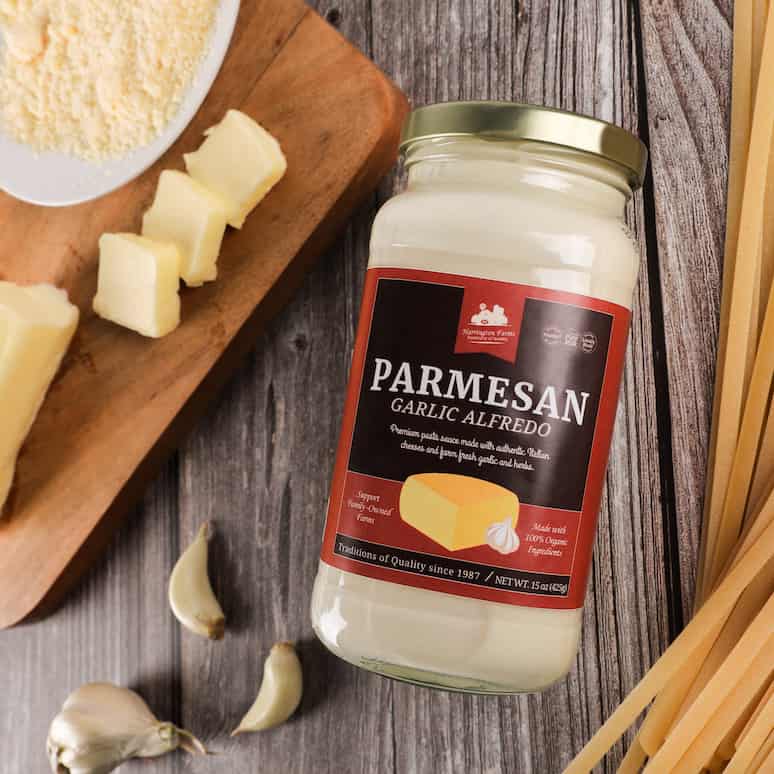 a jar of parmesan garlic alfredo sauce surrounded by dry fettucine noodles and garlic cloves, to the left of the jar is a cutting board with a knife, a partly sliced stick of butter and a bowl of dried parmesan cheeses