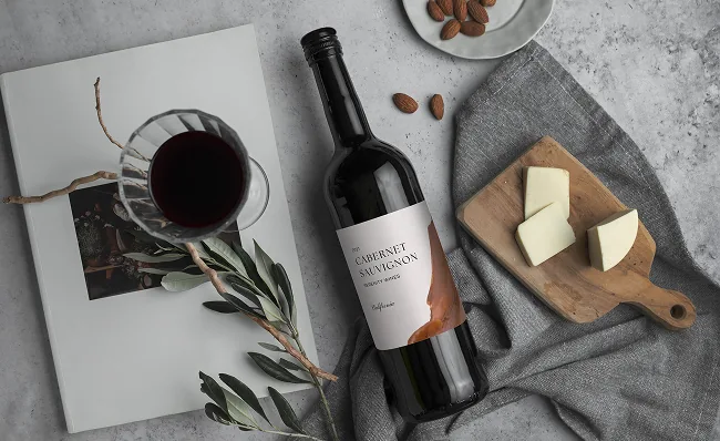 a glass of wine next to a bottle of red wine and a plate of nuts, next to the bottle is sliced cheese on a charcuterie board