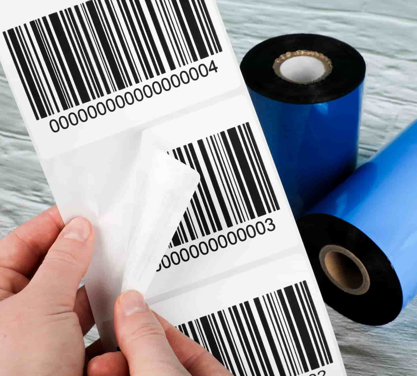 a hand peeling off a barcode label from a sheet with 3 in. core thermal ribbon rolls in the background