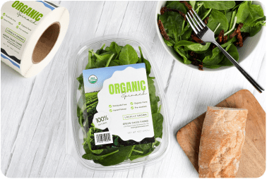 A plastic container of spinach with a high quality product label sitting next to fresh ingredients and a roll of those product labels