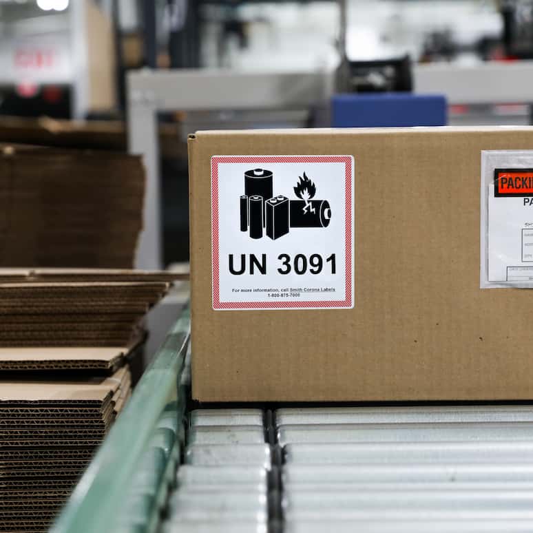 a UN 3091 label on a cardboard box, the box sits on a conveyor belt next to a stack of flat cardboard boxes