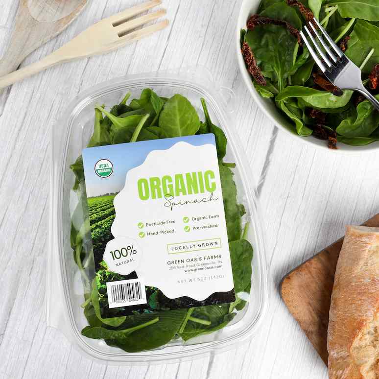 a package of spinach surrounded by a bowl of salad, half a baguette, and salad serving utensils