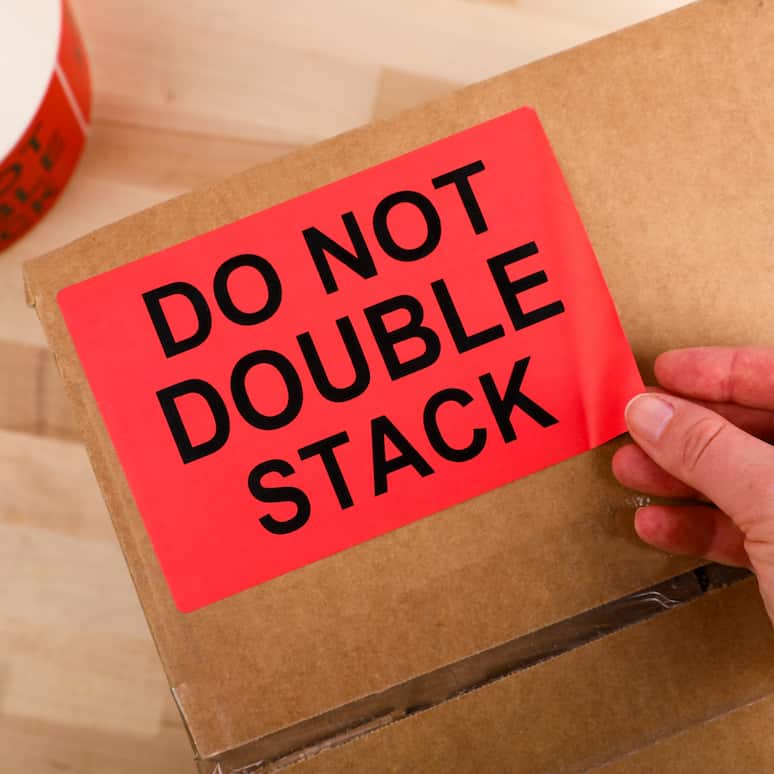 a hand putting a do not double stack label onto a cardboard box, on the floor is a roll of more labels