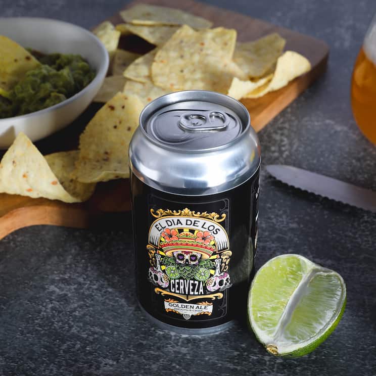 a can of golden ale on a counter surrounded by ships and guac, a halved lime next to a whole lime, a knife, and a glass of beer