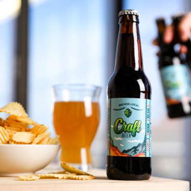 a bottle of craft beer on a table with a glass of beer, a bowl of chips. A hand is holding two more bottles in the background