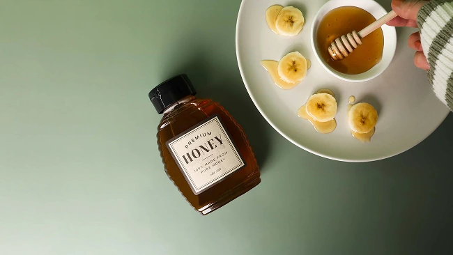 a bottle of honey sitting next to a plate, the plate holds a bowl of honey while a hand stirs it with a honey wand, bananas drizzled in honey flank the bowl in a radial pattern