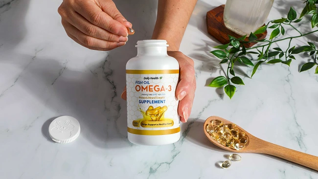 hands holding an omega-3 capsule and the other holding the pill bottle; a spoonful of the capsules sits to the right below a vine of fake foliage and a bottle of liquid