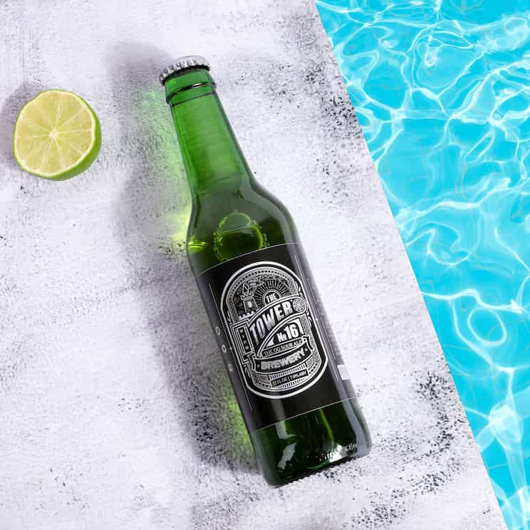 a bottle of beer on the pavement by a pool, to the left is a slice of lime