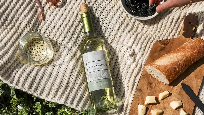 a bottle of white wine on a blanket to the right of a glass of the wine, to the right of the bottle is half a baguette next to some sliced cheese on a cutting board; a hand is picking up a blackberry from a bowl near the top of the frame