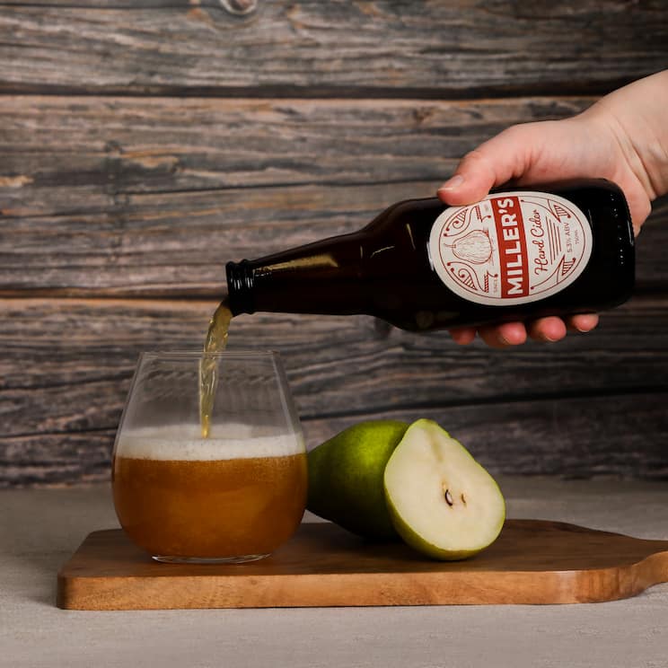 a hand pouring a bottle of hard cider into a glass next to a sliced pear