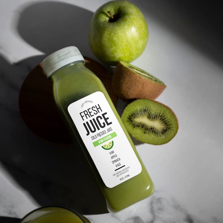 a bottle of kiwi juice sitting on a table among sliced kiwis and an apple, a glass of kiwi juice sits in the bottom left of the image