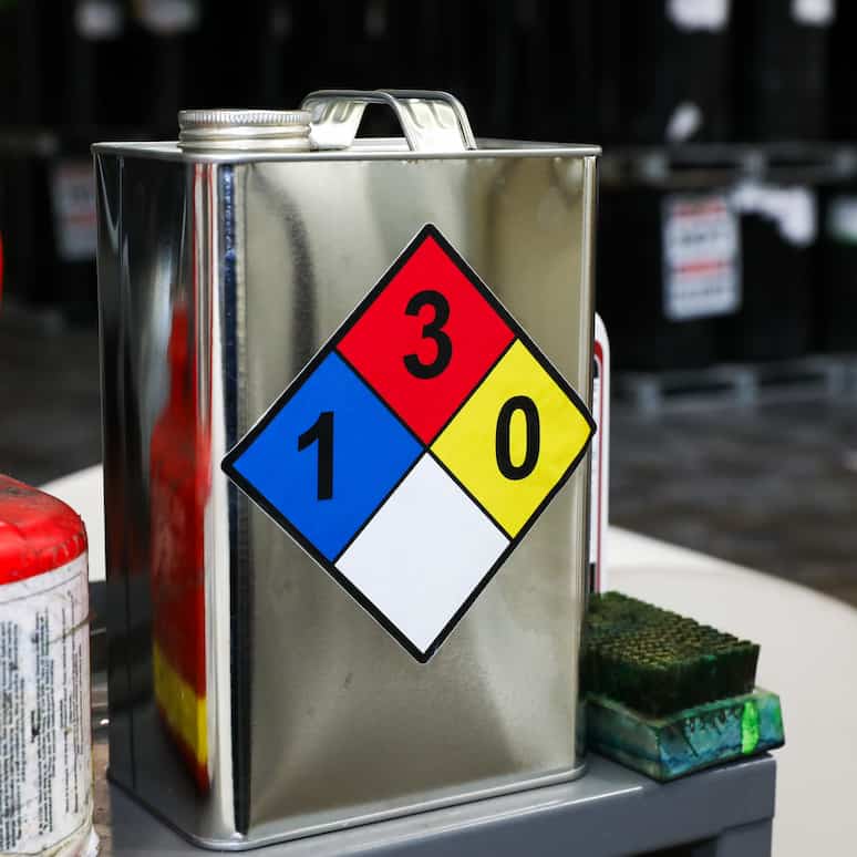 a metal jug with a NFPA diamond label on a metal table next to a metal container of ethanol, on the other side of the metal jug is a dirty scrub brush, in the background are stacked rows of large metal barrels