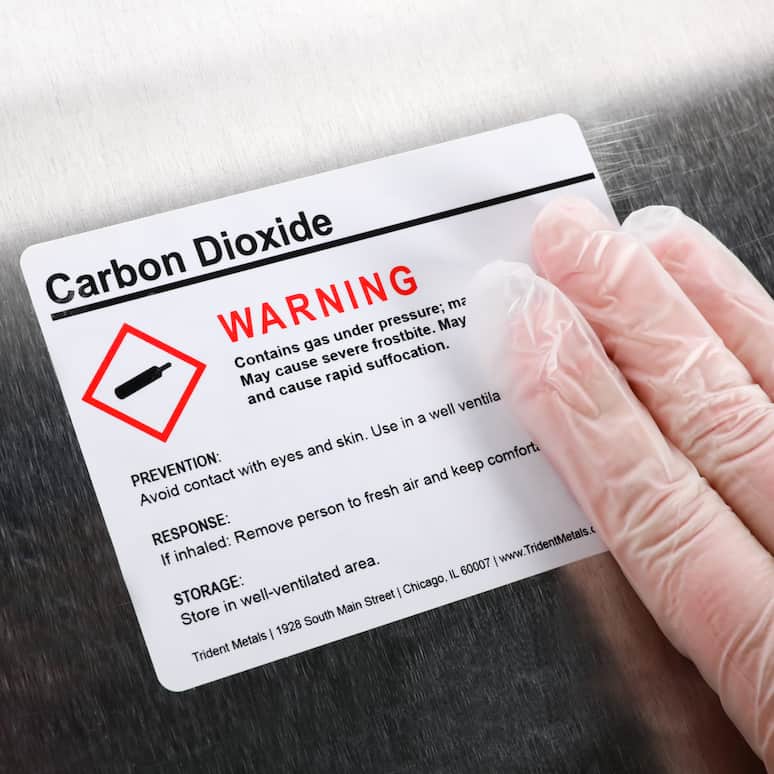 a hand wearing a plastic glove applies a label that has a warning about carbon dioxide