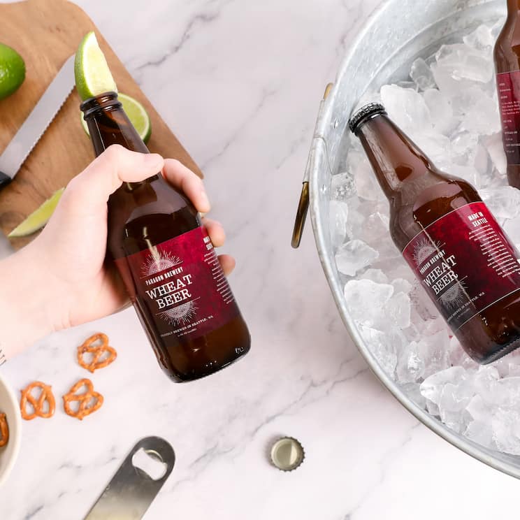 a hand holding a bottle of wheat beer with a slice of lime inserted in the top, in the background is a bucket of ice with 2 bottles of wheat beer, a cutting board with some limes, some pretzels, and a bottle opener