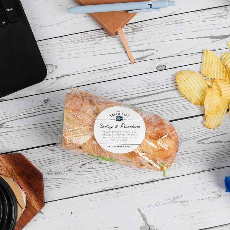 a saran wrapped turkey sandwich surrounded by a laptop, a notebook and pen, a handful of chips next to an open bag of potato chips, and a to-go cup of coffee on a wooden coaster