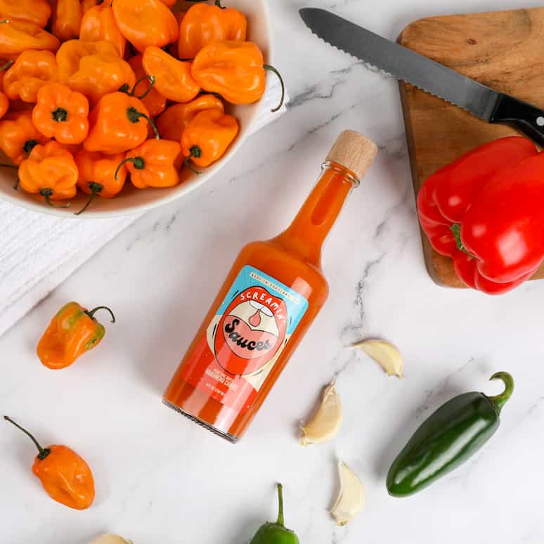 a bottle of Screamin Sauces hot sauce surrounded by habenaros, cloves of garlic, jalapenos, a red pepper and a serrated knife on a cutting board, and a bowl of habaneros