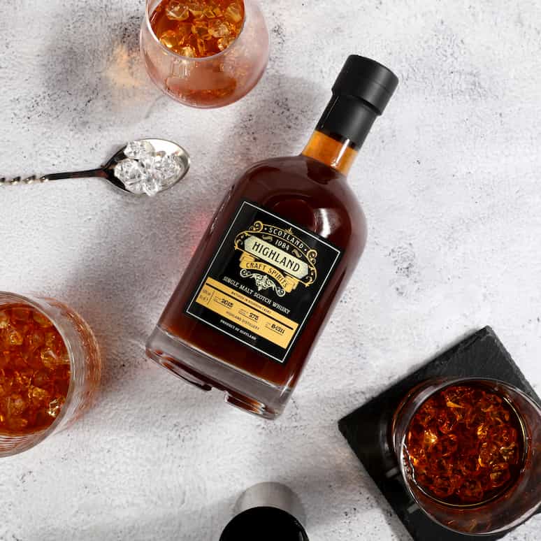 a bottle of bourbon surrounded by 3 glasses of bourbon over ice, a bar spoon holds some small ice cubes next to the bottle