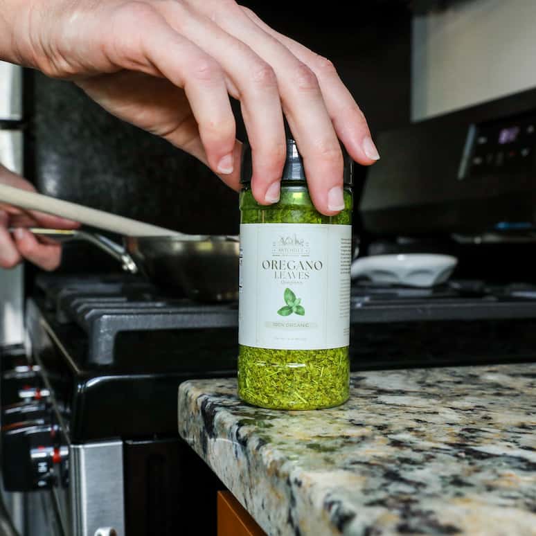 a hand reaching for a bottle of oregano on the counter while cooking food in a pan