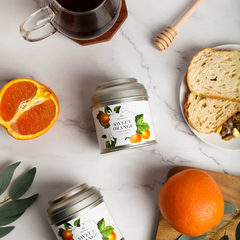 an overhead shot of two tins of sweet orange tea on a marble countertop, positioned around the tins are half an orange, some fake leaves, an orange sitting on a cutting board next to more leaves, some slices of bread and nuts on a plate, a honey wand, and a cup of tea on a wooden coaster