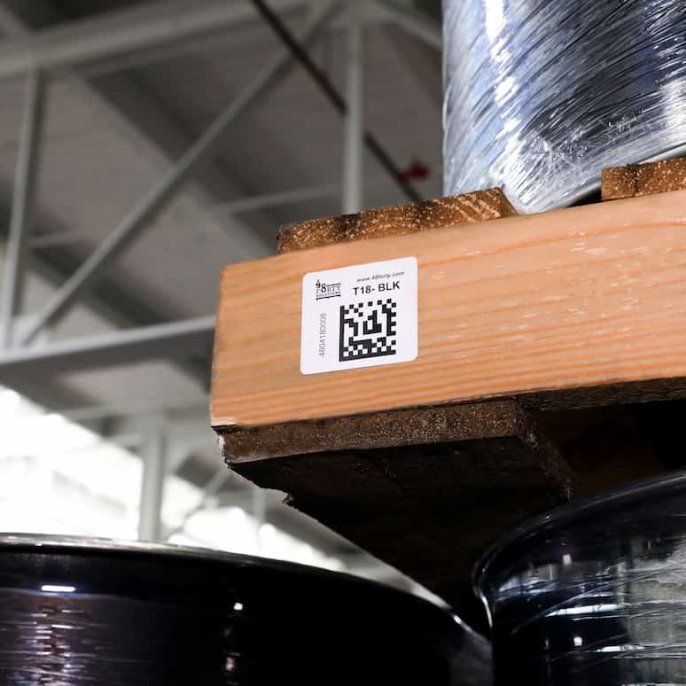 a data matrix label on a pallet sitting overhead, the shelves contain large plastic wrapped metal barrels