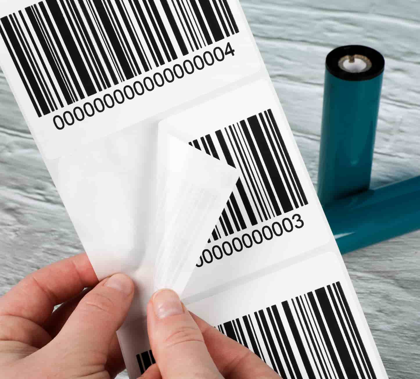 a hand peeling off a barcode label from a sheet with 2 thermal transfer ribbon rolls in the background