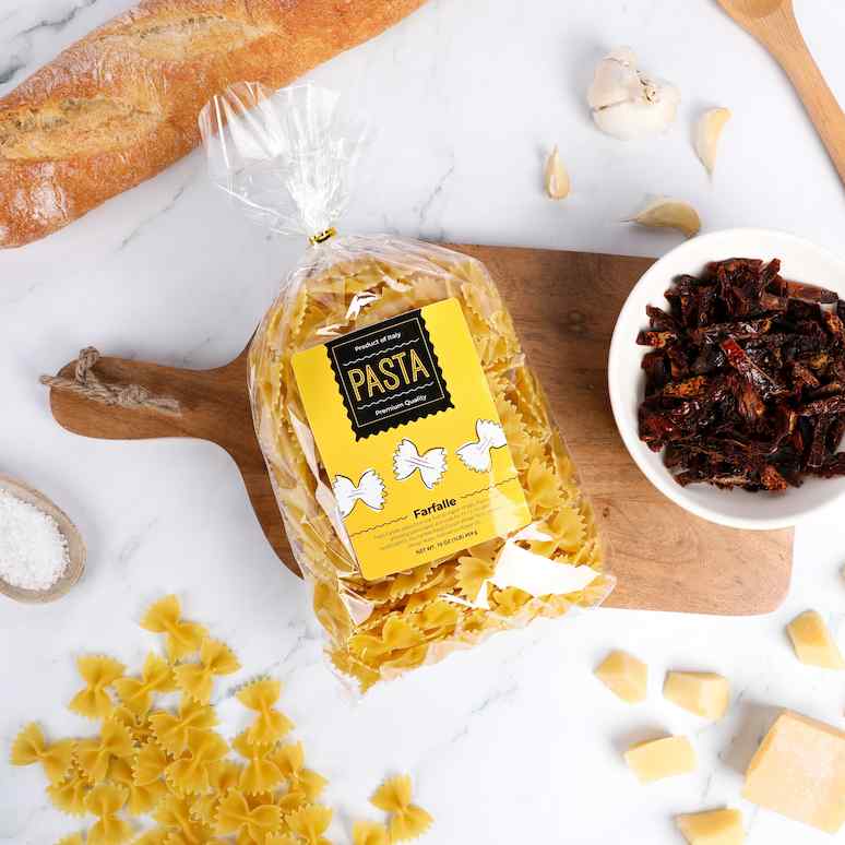 a labeled bag of pasta surrounded by a wooden spoon holding salt, a handful of bowtie pasta, a baguette, garlic cloves, dried red peppers, and sliced cheese