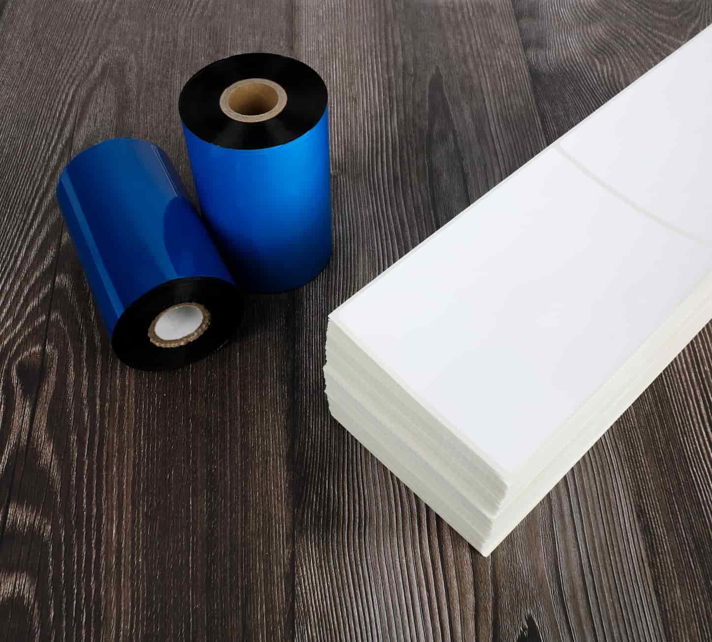 a stack of blank fanfold labels next to 2 thermal ribbons