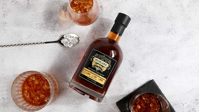 a top view of a bottle of whisky surrounded by glasses of whisky over ice and a bar spoon holding ice