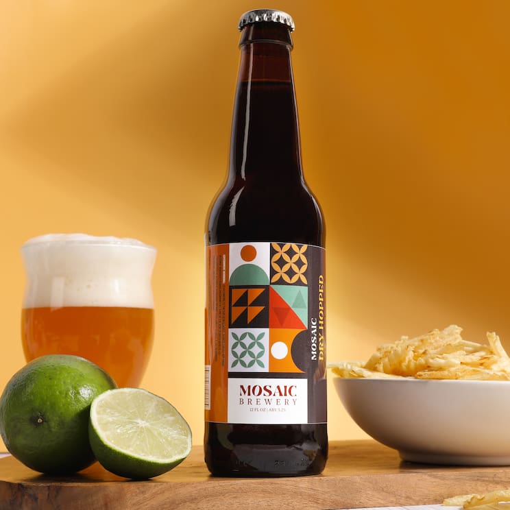a bottle of beer with a glass of beer and limes on the left and a bowl of chips on the right