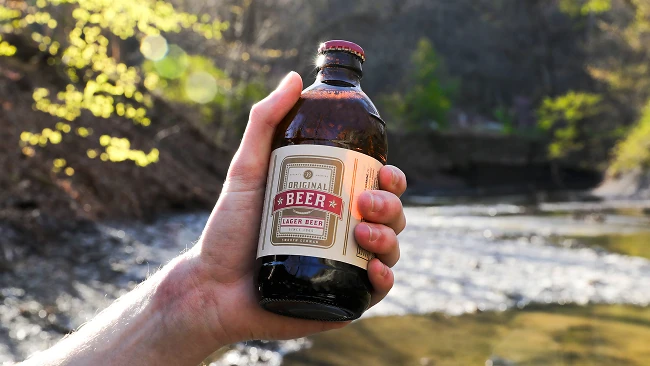a hand holding a bottle of lager near a stream