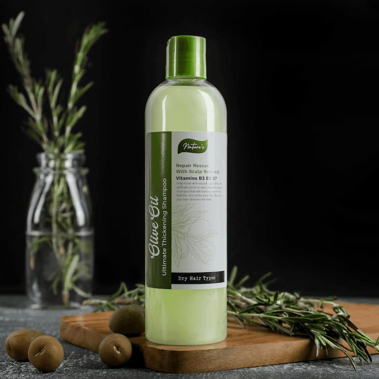 a bottle of olive oil shampoo sitting on a cutting board next to rosemary and a few scattered olives, in the backround is more rosemary sitting in a milk bottle half full with water