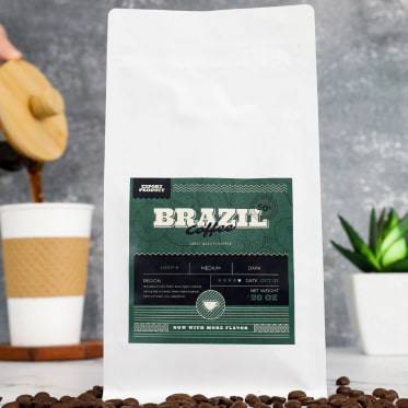 a labeled bag of Brazilian coffee, directly in front are a bunch of spilled coffee beans, behind the bag to the left is a pot of coffee being poured into a to-go cup, to the right is a potted plant