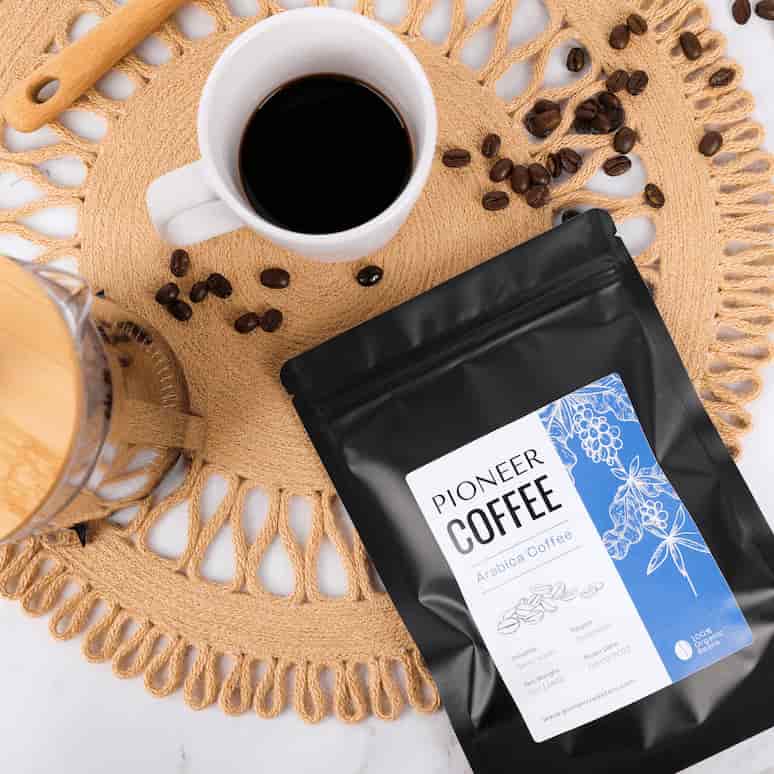 an overhead view of a labeled bag of coffee on a table with spilled coffee beans, a cup of coffee and a french press